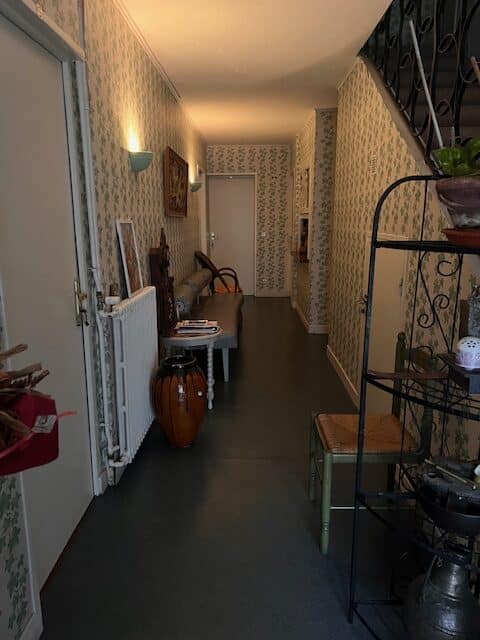Couloir d'entrée traditionnel. Papier peint feuillage vert, sol sombre, appliques lumineuses. Banc, radiateur et grande vase décorative près de l'escalier.
