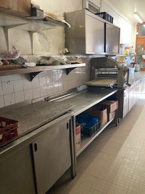 Cuisine professionnelle avec laminoir à pâte sur plan de travail en inox et étagères de rangement.