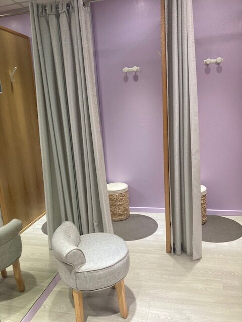 Cabines d'essayage aux murs violets, rideaux gris, tabourets en osier et chaise grise dans une boutique moderne.