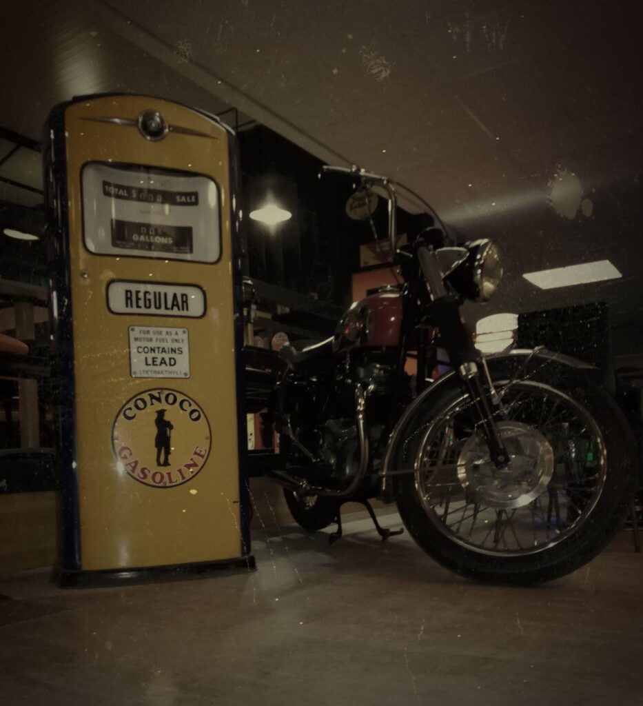 Pompe vintage Conoco jaune et moto de collection dans un garage rétro. Texte visible: Conoco Gasoline et Regular.