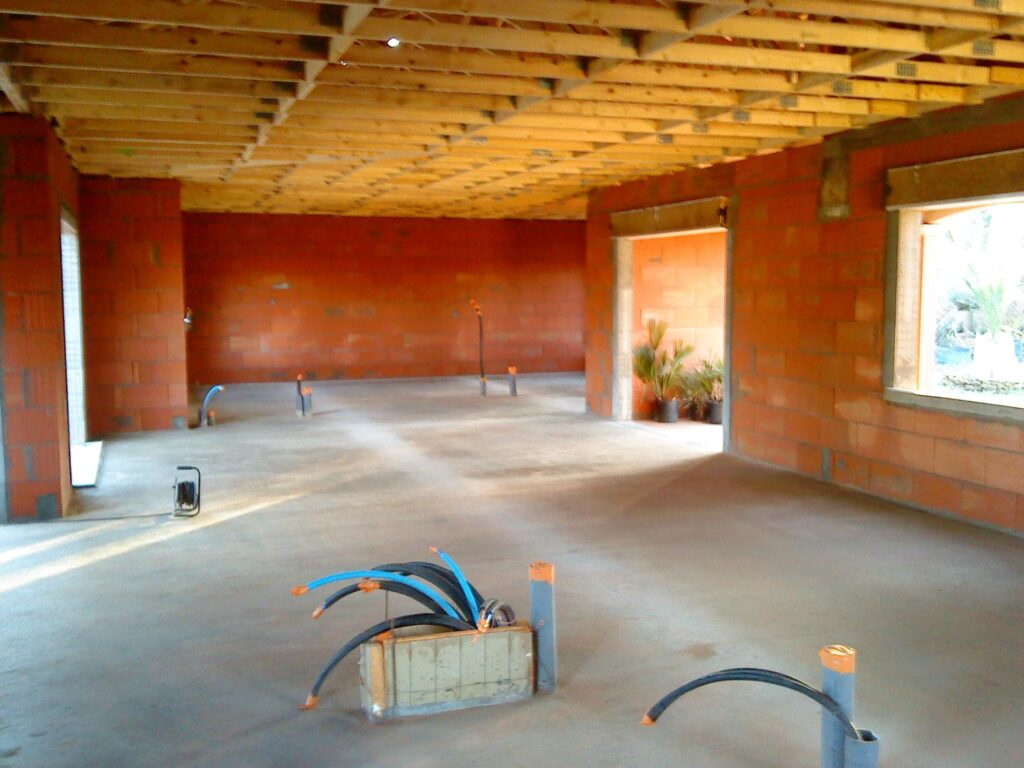 Intérieur de maison en construction avec murs en briques rouges, plafond en bois et conduits électriques au sol.