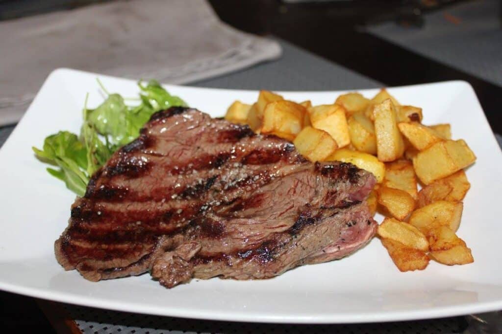 Steak grillé juteux, pommes de terre sautées et salade verte sur une assiette blanche.