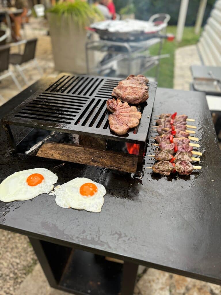 Oeufs au plat, steaks et brochettes grillant sur un brasero plancha au feu de bois dans un jardin.