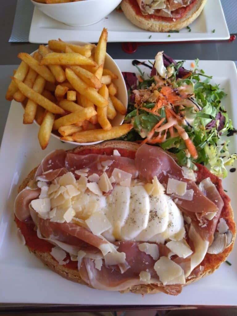 Tartine au jambon cru, mozzarella et parmesan avec frites dorées et salade fraîche sur une assiette blanche.