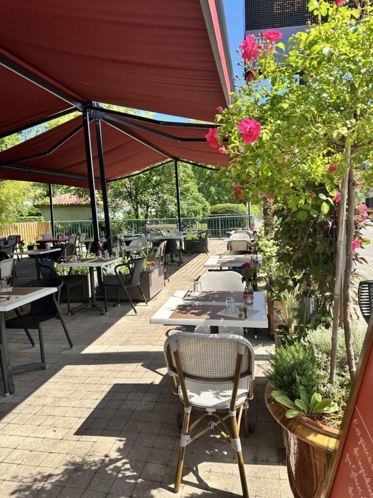 Terrasse de restaurant sous stores rouges avec tables et rosiers roses.