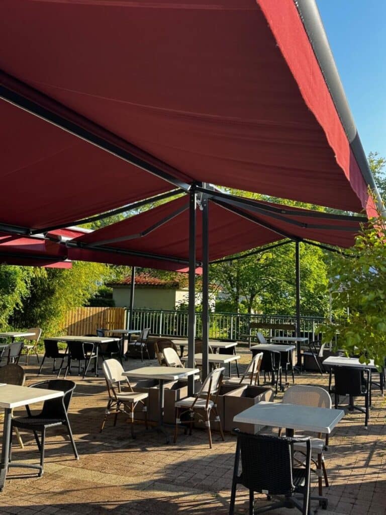 Terrasse de restaurant ombragée par des stores rouges avec tables blanches et chaises modernes.