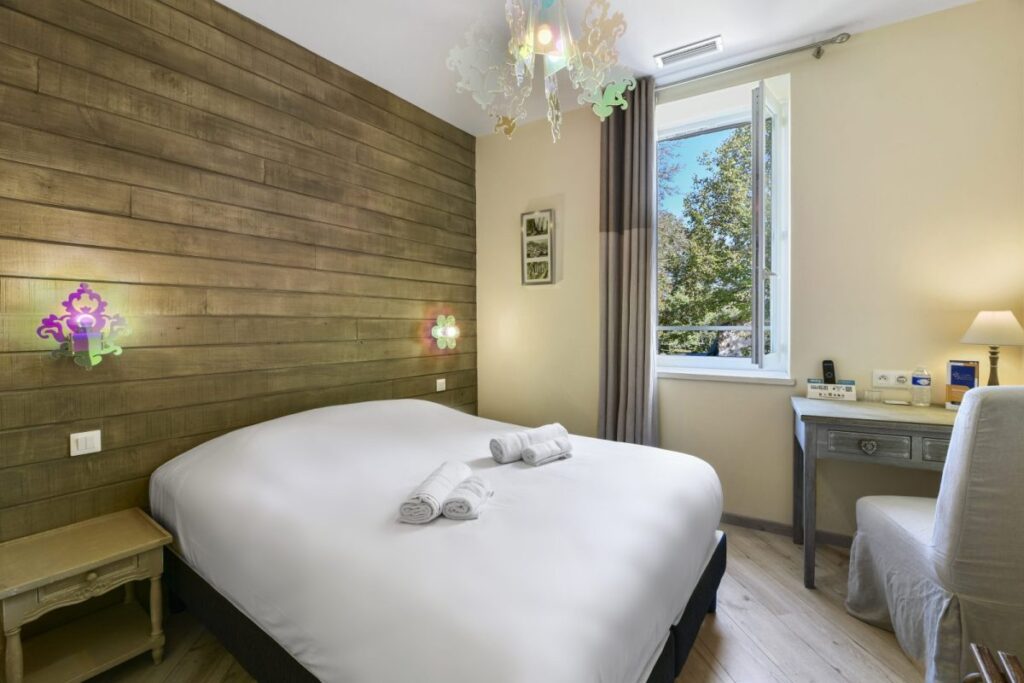 Chambre d'hôtel lumineuse avec lit double, mur boisé, bureau et luminaire design devant une fenêtre ouverte.