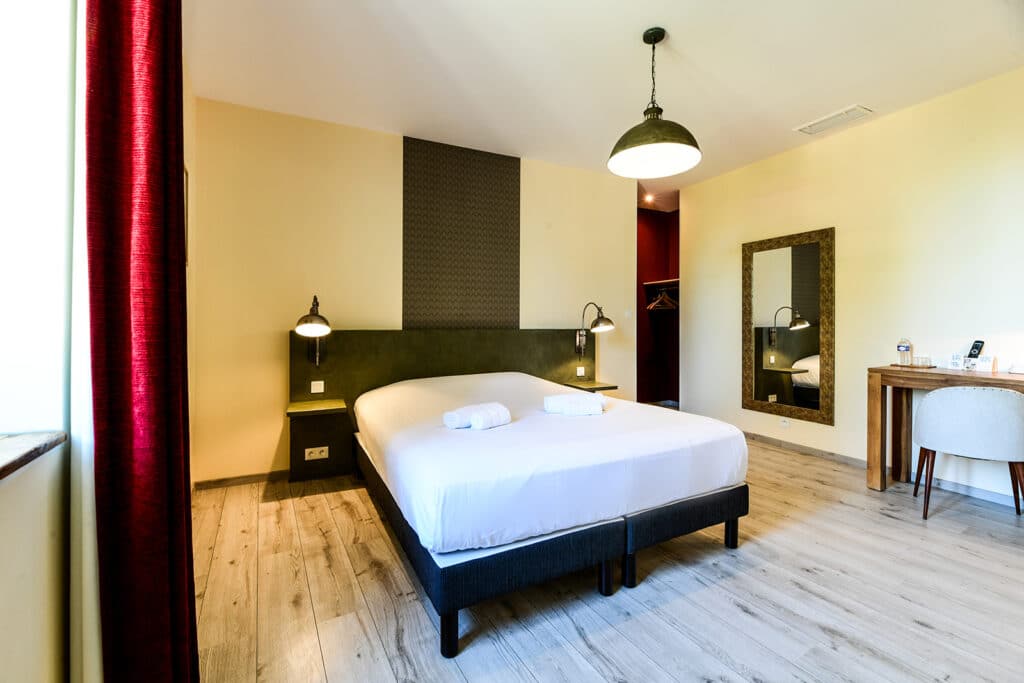 Chambre d'hôtel moderne avec grand lit double, parquet clair, bureau, miroir et rideaux rouges élégants.