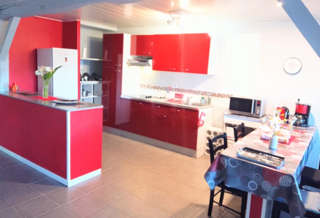 Cuisine moderne rouge et blanche avec bar, électroménagers, table à manger dressée et carrelage clair au sol.