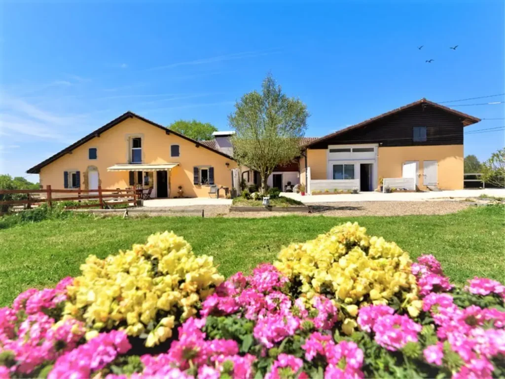 Ferme jaune avec jardin aux fleurs roses et jaunes sous un ciel bleu, maison de campagne paisible en été.