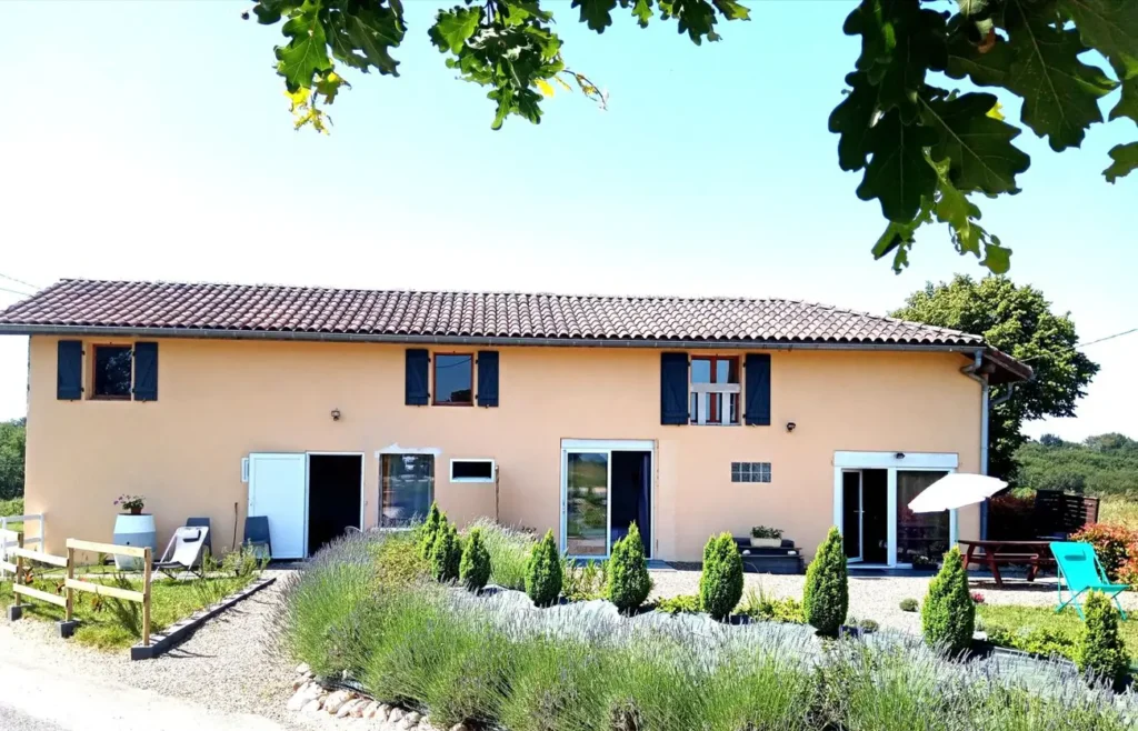 Longue maison orange aux volets bleus avec jardin de lavande et petits conifères décoratifs.