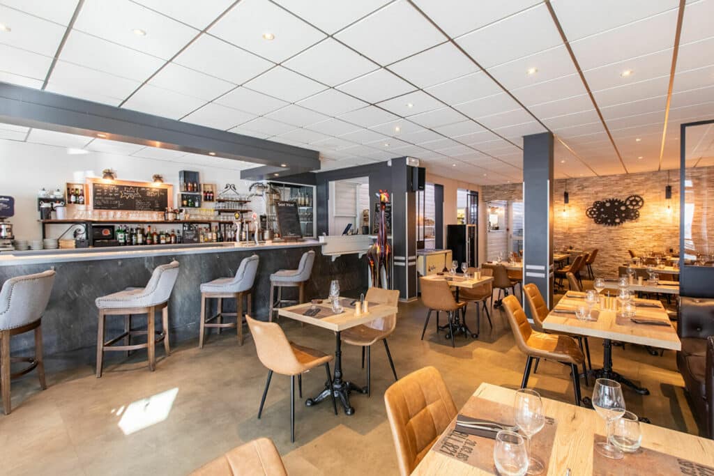 Salle de restaurant moderne avec bar gris, chaises en cuir et tables en bois sous une ambiance lumineuse chaleureuse.