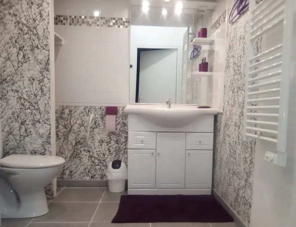 Salle de bain avec papier peint motif branches, meuble vasque blanc, tapis prune et radiateur sèche-serviettes.