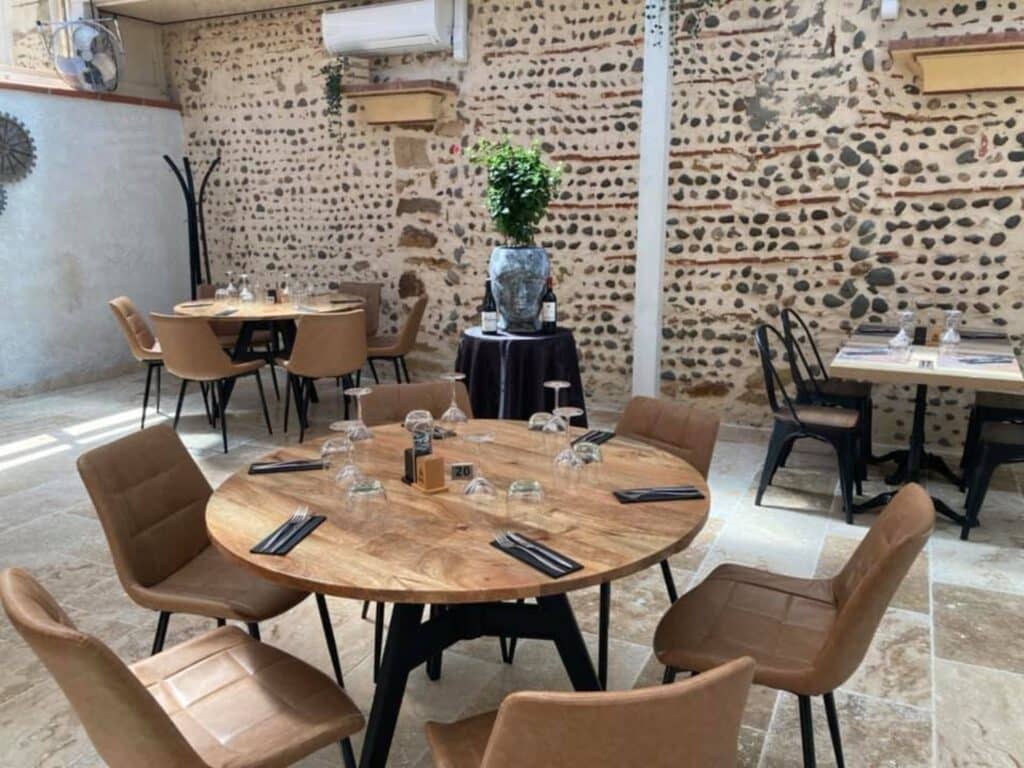 Salle de restaurant rustique : murs en pierre, tables en bois et chaises marron. Table 20 dressée avec verres.
