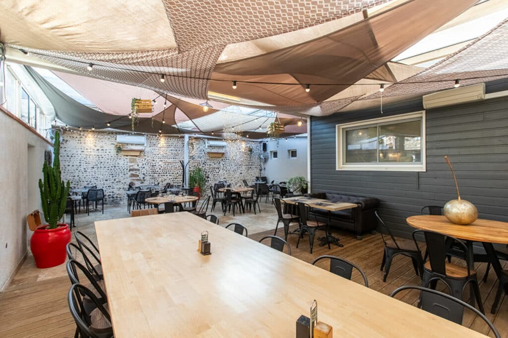 Terrasse design : voiles d'ombrage, guirlandes, tables bois et chaises industrielles devant un mur en pierre.