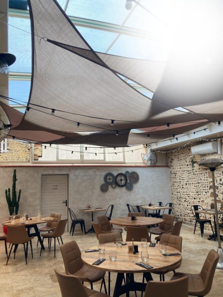 Terrasse de restaurant avec voiles d'ombrage, tables en bois rondes et murs en pierre au style industriel.